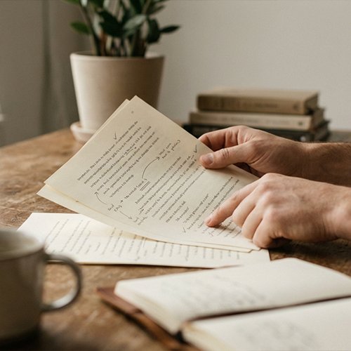 Person reviewing marked and corrected papers, with a cup and notebook on the table.