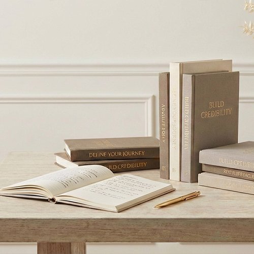 Stack of books with titles 'Build Credibility' and 'Define Your Journey', along with an open notebook and pen on a wooden table